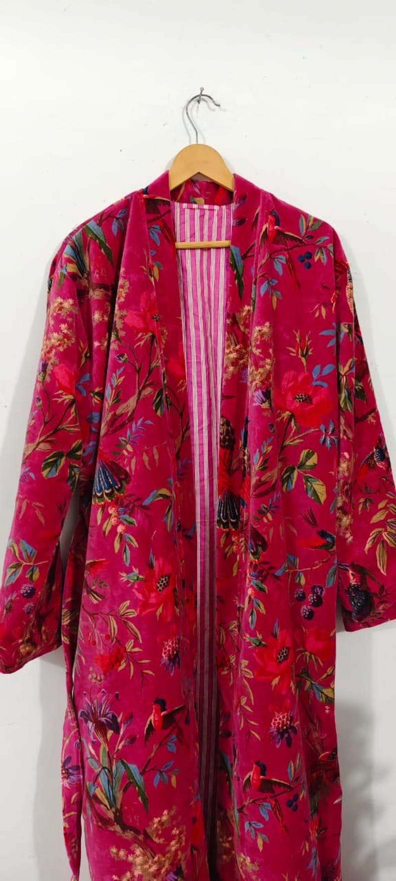 Puede incluir: Bata larga y abierta de color magenta vibrante, adornada con un estampado floral y de p&aacute;jaros en tonos rojos, verdes, azules y dorados. La bata presenta un forro interior a rayas y se exhibe en una percha de madera.