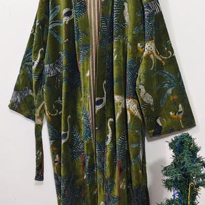 May include: A long, olive green velvet robe with a jungle print featuring leopards, birds, and palm trees. The robe has a striped pink and white lining and a matching belt. It is hanging on a wooden hanger.