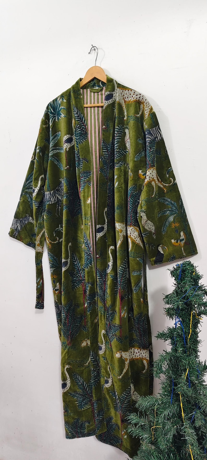 May include: A long, olive green velvet robe with a jungle print featuring leopards, birds, and palm trees. The robe has a striped pink and white lining and a matching belt. It is hanging on a wooden hanger.