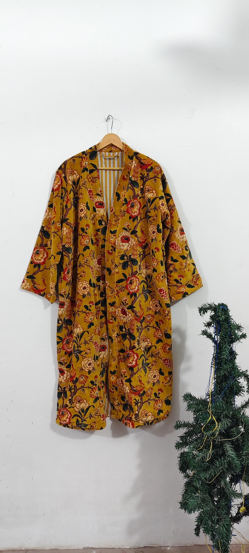 Puede incluir: Bata estilo kimono con estampado floral amarillo mostaza. La bata presenta un estampado floral a gran escala en rojo, naranja y verde. El forro interior tiene un estampado de rayas. La bata est&aacute; colgada de una percha de madera.