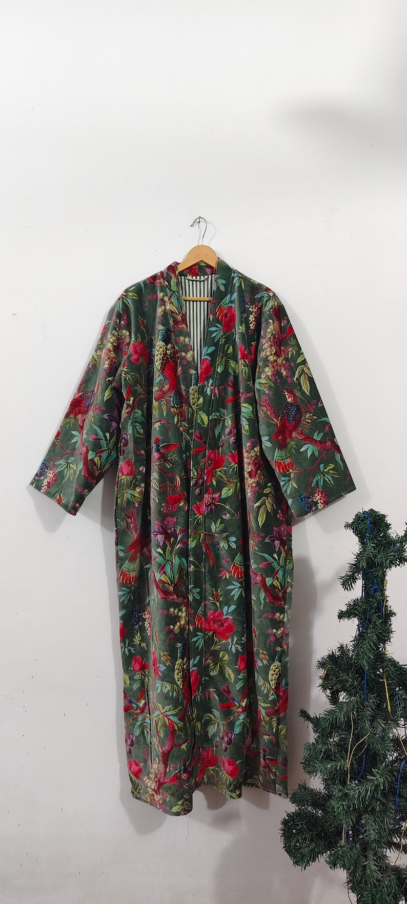 Puede incluir: Larga bata de terciopelo verde con un estampado floral y de p&aacute;jaros. La bata tiene un forro a rayas y est&aacute; colgada de una percha de madera. El estampado floral incluye flores rojas y moradas.