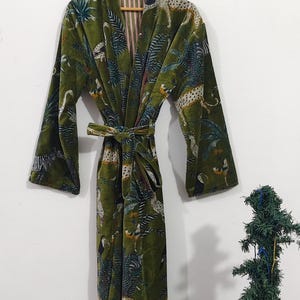 May include: A long, olive green velvet robe with a tie belt, hanging on a wooden hanger. The robe features a jungle-themed pattern with leopards, toucans, and tropical plants. The interior lining has a striped pattern.