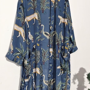 May include: A blue kimono-style robe with a jungle-themed pattern. The robe features a repeating design of leopards, birds, and palm trees in shades of white, yellow, and green. The robe is hanging on a wooden hanger.