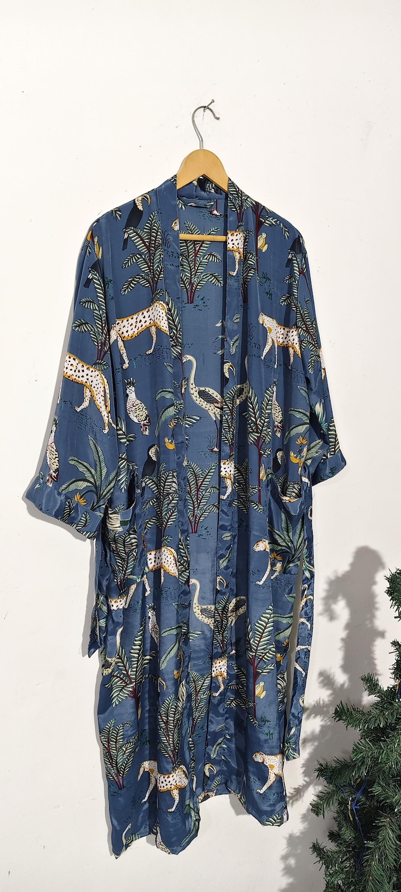 May include: A blue kimono-style robe with a jungle-themed pattern. The robe features a repeating design of leopards, birds, and palm trees in shades of white, yellow, and green. The robe is hanging on a wooden hanger.