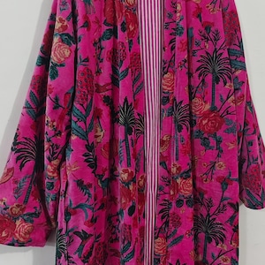 Puede incluir: Una bata rosa vibrante con un estampado floral y de palmeras. La bata tiene un interior a rayas y est&aacute; colgada de una percha de madera. El dise&ntilde;o incluye detalles en rojo, verde y negro.