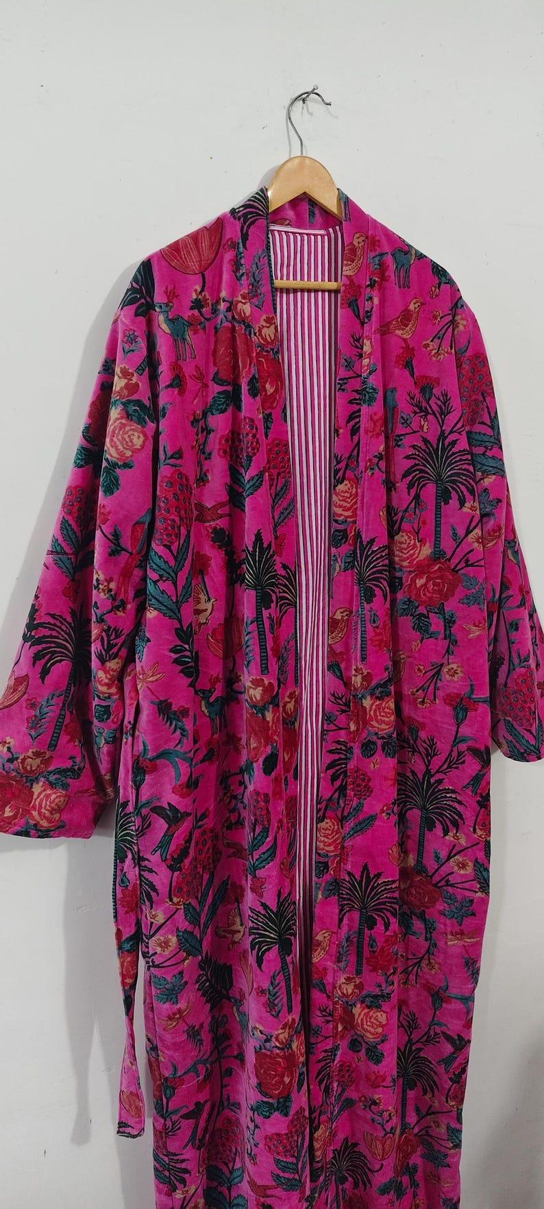 Puede incluir: Una bata rosa vibrante con un estampado floral y de palmeras. La bata tiene un interior a rayas y est&aacute; colgada de una percha de madera. El dise&ntilde;o incluye detalles en rojo, verde y negro.