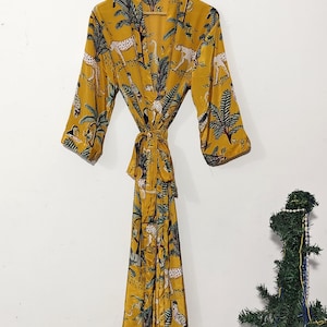 May include: Mustard yellow robe with a jungle print featuring leopards and palm trees. The robe has long sleeves and a matching tie belt. The robe is hanging on a wooden hanger against a white background.