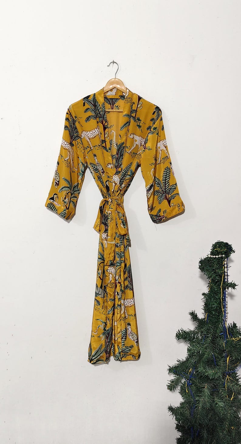 May include: Mustard yellow robe with a jungle print featuring leopards and palm trees. The robe has long sleeves and a matching tie belt. The robe is hanging on a wooden hanger against a white background.