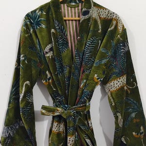 Puede incluir: Bata de terciopelo verde oliva con estampado tropical con leopardos, p&aacute;jaros y palmeras. La bata tiene un cintur&oacute;n a juego y forro a rayas. La bata est&aacute; colgada de una percha de madera.