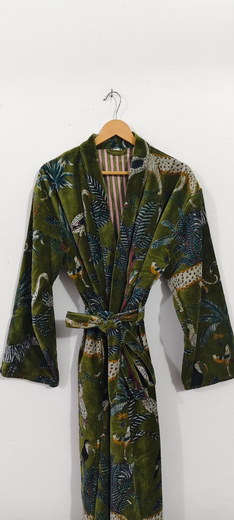 Puede incluir: Bata de terciopelo verde oliva con estampado tropical con leopardos, p&aacute;jaros y palmeras. La bata tiene un cintur&oacute;n a juego y forro a rayas. La bata est&aacute; colgada de una percha de madera.
