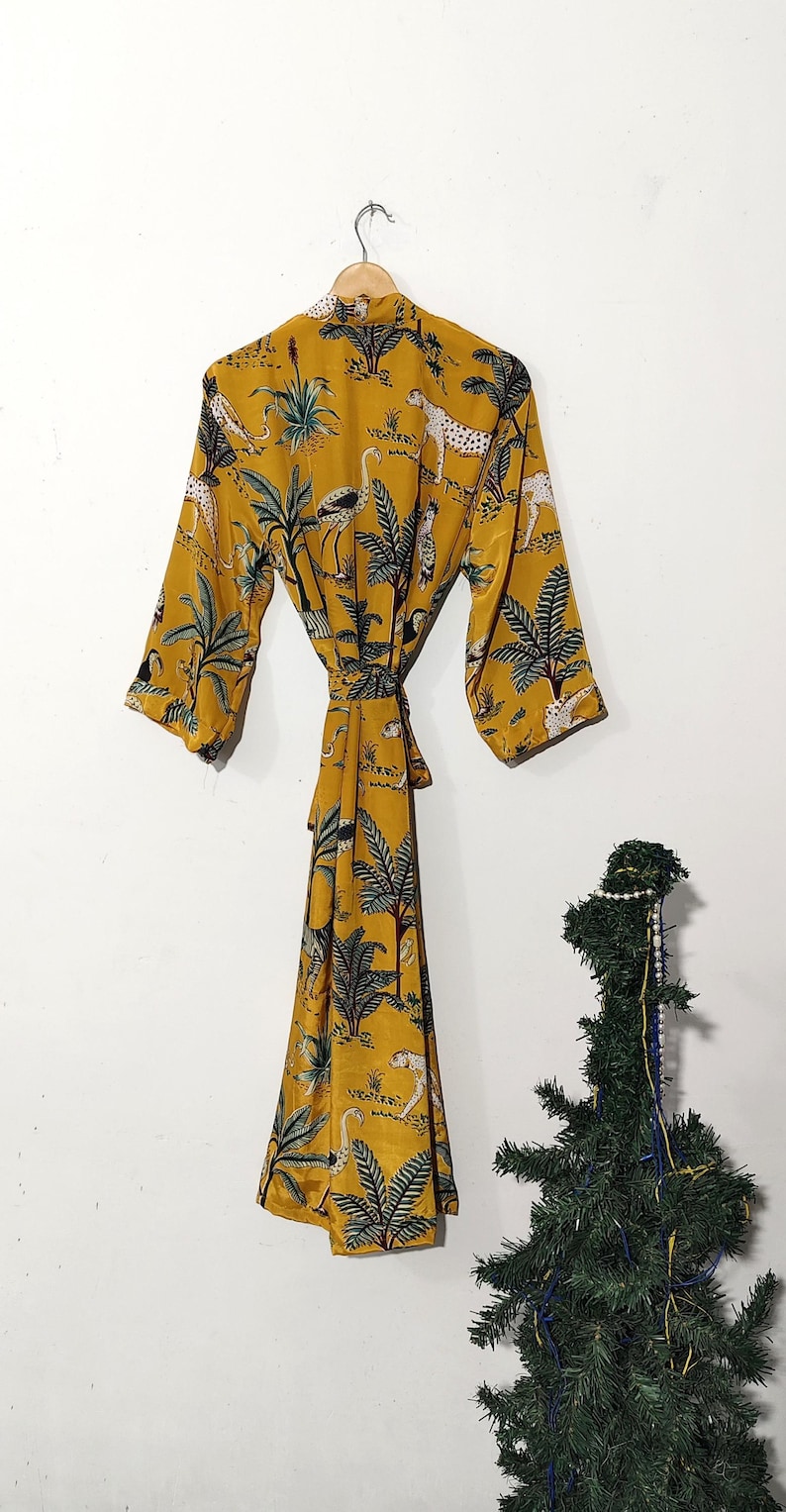 May include: Mustard yellow robe with a jungle print featuring leopards, birds, and palm trees. The robe has a tie waist and three-quarter sleeves. The robe is hanging on a wooden hanger.