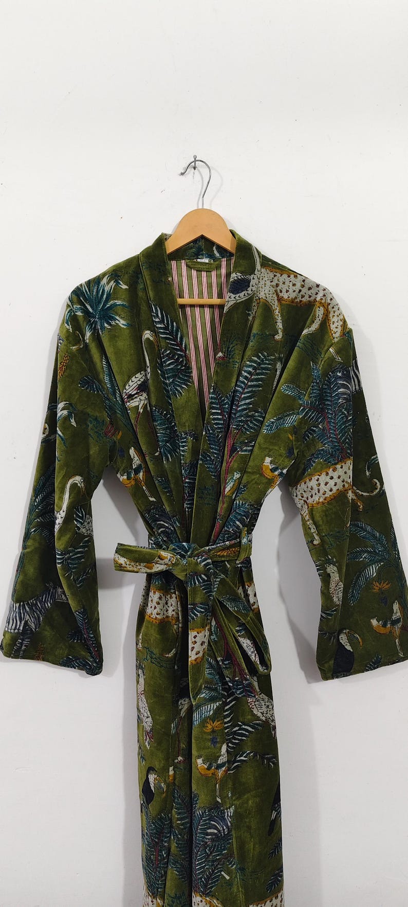 May include: A green velvet robe with a tropical pattern featuring leopards, birds, and palm trees. The robe has a matching belt and a striped lining. The robe is hanging on a wooden hanger.