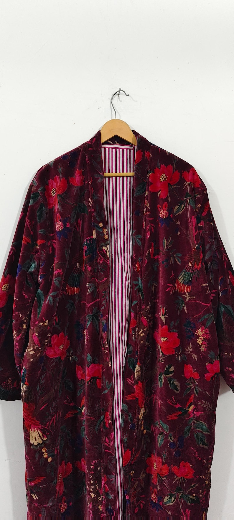 Puede incluir: Kimono de terciopelo burdeos con un vibrante estampado floral en rojo, verde y azul. El forro interior presenta rayas verticales blancas y moradas. Esta bata abierta cuelga de una percha de madera.