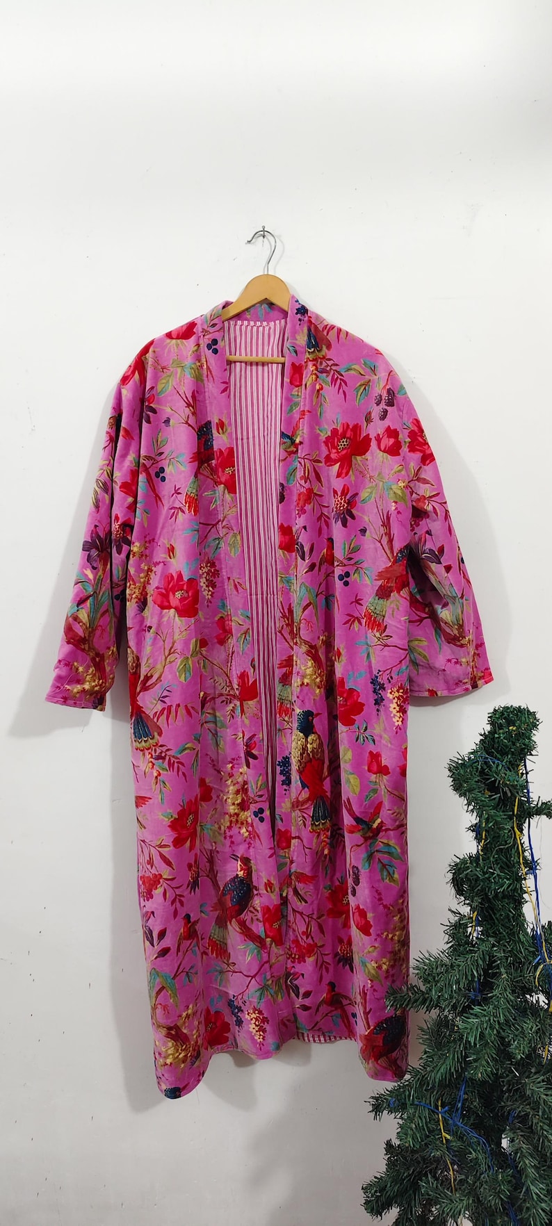 Puede incluir: Una bata larga y abierta de color rosa vibrante, adornada con un estampado floral y de p&aacute;jaros en rojo, verde y azul. El interior de la bata presenta una tela a rayas rosas y blancas. La prenda est&aacute; colgada de una percha de madera.