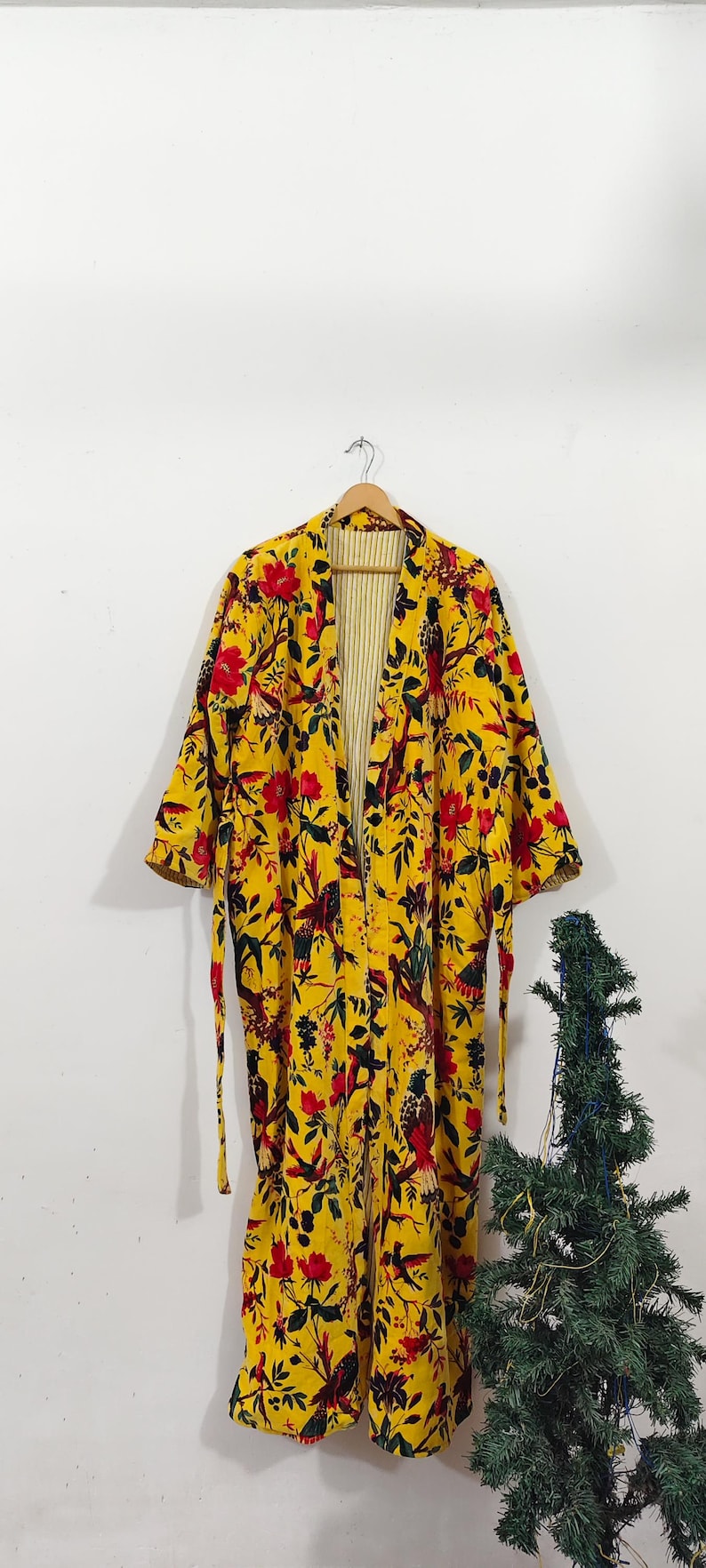 Puede incluir: Larga bata kimono amarilla con estampado floral, flores rojas y detalles de p&aacute;jaros y hojas negras. La bata tiene un interior a rayas y est&aacute; colgada de una percha de madera. La bata tiene mangas largas y cintur&oacute;n anudado.