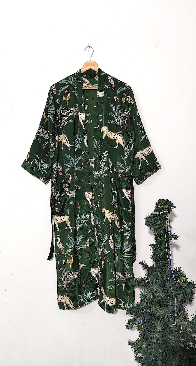 May include: A dark green kimono-style robe with a jungle-themed pattern. The robe features a repeating design of leopards, zebras, and tropical foliage. The robe is hanging on a wooden hanger.