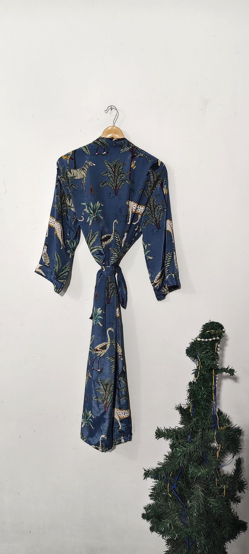 May include: A blue robe with a jungle animal print, including zebras, leopards, and birds, hangs on a wooden hanger. The robe is tied at the waist with a matching fabric belt. The robe is made of a silky material.