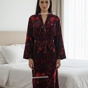 Puede incluir: Bata de terciopelo burdeos con estampado floral. La bata tiene mangas largas, cintur&oacute;n anudado y dobladillo hasta la rodilla. El dise&ntilde;o floral incluye detalles en rojo y verde. La bata se muestra en un dormitorio.