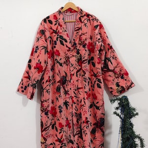 Puede incluir: Bata larga con estampado floral de flores rojas y hojas verde oscuro sobre fondo rosa. La bata est&aacute; colgada de una percha de madera. El forro interior es a rayas.