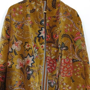 May include: Mustard-colored velvet jacket with a floral pattern in pink, red, and blue. The open front jacket has a striped lining in brown and white. The jacket is hanging on a wooden hanger against a white background.