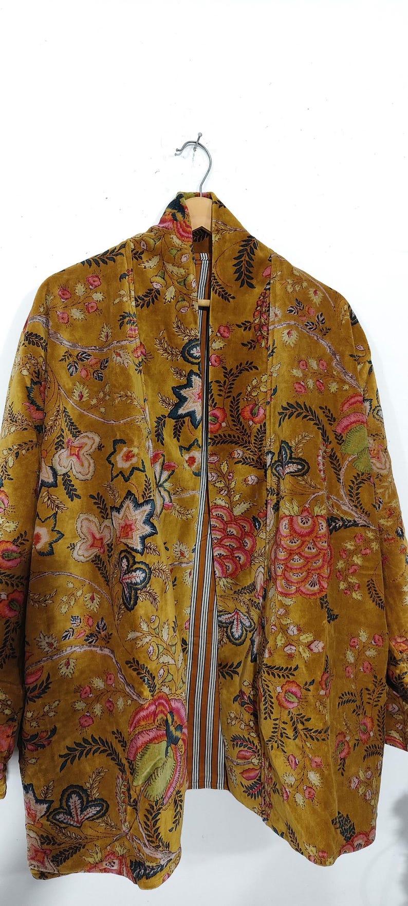May include: Mustard-colored velvet jacket with a floral pattern in pink, red, and blue. The open front jacket has a striped lining in brown and white. The jacket is hanging on a wooden hanger against a white background.