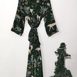 May include: A dark green robe with a jungle and leopard print. The robe is hanging on a wooden hanger against a white wall. The robe has long sleeves and a matching tie belt.