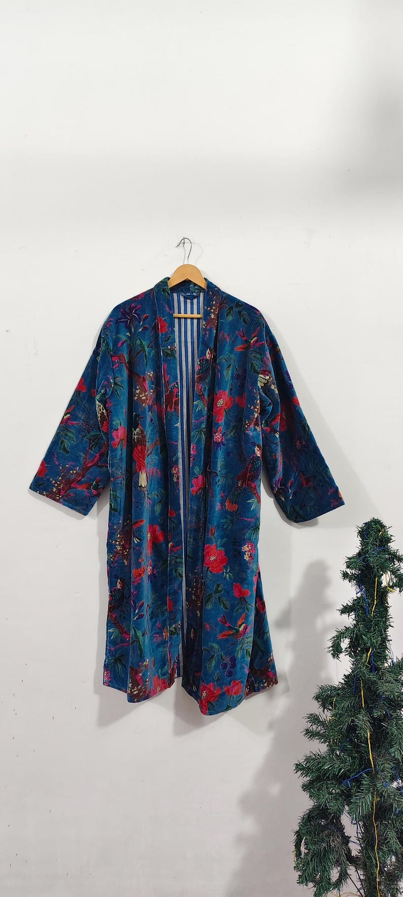 May include: A long, open-front robe in a teal blue hue, adorned with a vibrant floral pattern in red and green. The robe features long sleeves and a striped inner lining. It is hanging on a wooden hanger.