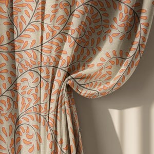 May include: Close-up of a beige curtain with a repeating pattern of orange leaf-like shapes and dark gray stems. The curtain is draped, revealing the texture and design. The background is a neutral wall with shadows.