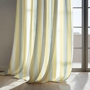 May include: Yellow and white striped curtains with blue dotted stripes. The curtains are hanging in front of a window, and the fabric has a textured appearance. The curtains are made of a light, flowing material.