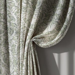 May include: Curtains with a repeating pattern of green leaves on a cream background. The fabric is draped, showing folds and texture. The curtains are hanging against a neutral gray wall, with shadows.