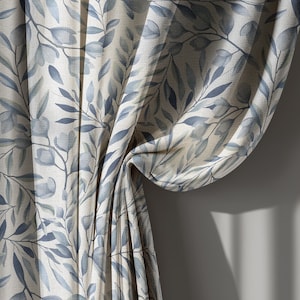 May include: Curtains with a botanical pattern. The fabric features a repeating design of blue-grey leaves and floral elements on a cream background. The curtains are draped, showing folds and texture, with shadows cast on the wall.