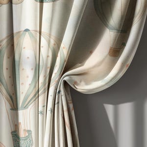 May include: Curtains with a repeating pattern of watercolor hot air balloons in muted tones of beige, blue, and peach. The fabric is textured and draped, with visible folds and shadows. The design evokes a whimsical, dreamy aesthetic.