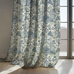 May include: Blue and cream-colored floral patterned curtain panels. The fabric features a detailed design of flowers, foliage, and medallion-style motifs. The curtains are hanging in front of a window, with natural light illuminating the fabric.