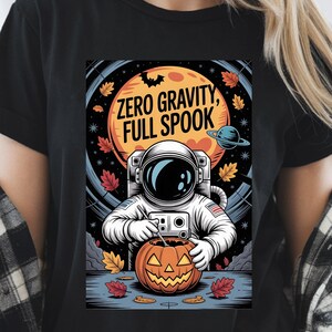 May include: Black t-shirt with a Halloween-themed graphic. The design features an astronaut carving a jack-o'-lantern with the text "ZERO GRAVITY, FULL SPOOK" above. The background includes a full moon, planets, and autumn leaves.