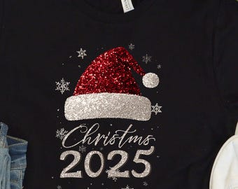 Camiseta de Navidad 2025 con gorro rojo de Papá Noel y diseño de copo de nieve brillante para celebraciones festivas.