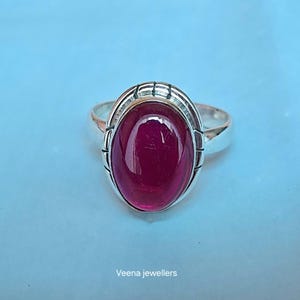 Può includere: Un anello d'argento con una gemma ovale rosso scuro. La gemma è incastonata in una lunetta decorativa con un design strutturato. L'anello è su uno sfondo azzurro. Il testo "Veena jewellers" è in basso.