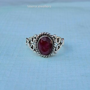Puede incluir: Un anillo de plata con una piedra preciosa roja oscura ovalada. La piedra preciosa está rodeada por un diseño de cuerda trenzada y detalles de filigrana ornamentados. El anillo está sobre un fondo azul claro. El texto "Veena jewellers" está arriba a la izquierda.