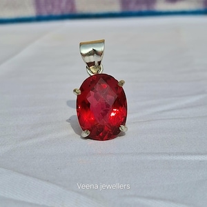 Op de afbeelding: Zilveren hanger met een grote, facetgeslepen, ovaalvormige rode edelsteen. De edelsteen wordt op zijn plaats gehouden door vier kleine pootjes. De hanger heeft een zilveren bale aan de bovenkant. De tekst "Veena jewellers" staat onderaan.