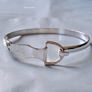 Könnte beinhalten: Ein silberfarbenes Armreif-Armband mit einem einzigartigen Verschlussdesign. Das Armband hat ein abgerundetes Band und einen Verschluss, der einem Steigbügel ähnelt. Der Markenname "Veena jewellers" ist oben links sichtbar.