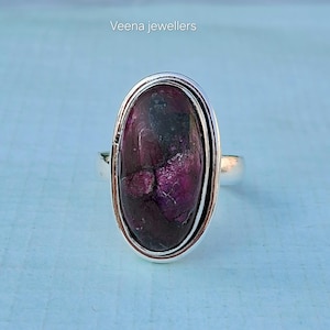 May include: A silver ring featuring an oval-shaped, deep purple gemstone. The stone has a mottled appearance with lighter purple and black inclusions. The ring's band is a simple, polished silver. The text "Veena jewellers" is visible in the upper left corner.