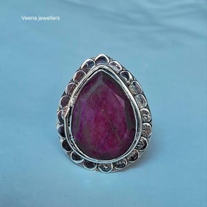 Può includere: Un anello a forma di goccia con una grande pietra preziosa sfaccettata rosa scuro incastonata in una fascia d'argento. L'incastonatura è circondata da un bordo decorativo smerlato con finitura testurizzata. Il testo Veena jewellers è visibile.