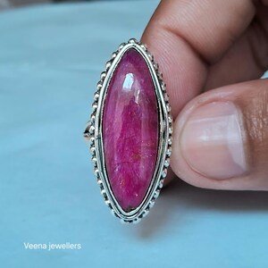 Puede incluir: Un anillo en forma de marquesa con una gran piedra preciosa de color rosa vibrante engastada en un marco de plata pulida con un borde de cuentas. La piedra muestra una textura fibrosa natural en su superficie. El texto Veena jewellers es visible abajo.
