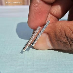 May include: Close-up of two silver bar earrings. The earrings are held between fingers against a light blue background. The earrings are rectangular and have a polished finish. The text "Veena jewellers" is in the top left corner.