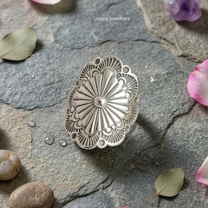 Peut inclure: Une bague statement argentée avec un motif floral détaillé. La bague présente un motif central en forme d'éventail entouré de bords festonnés. Elle est présentée sur une surface en pierre grise avec des feuilles et des pétales éparpillés.
