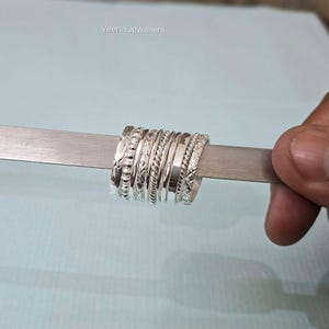 Könnte beinhalten: Eine Sammlung von Silberringen mit verschiedenen strukturierten Designs, darunter Perlen-, geflochtene und gemusterte Bänder. Die Ringe werden auf einem silbernen Messwerkzeug vor einem hellblauen Hintergrund präsentiert. Die Ringe werden von einer Hand gehalten.