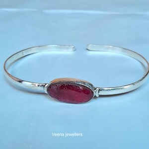 May include: A silver cuff bracelet featuring a large, oval, deep red gemstone. The bracelet has an open design and a polished finish. The background is a light blue colour.