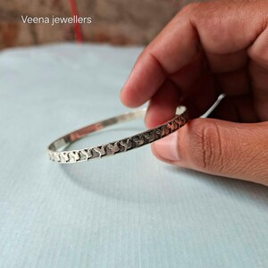 May include: A silver bangle with an engraved pattern of repeating shapes. The bangle is held in a hand against a light blue background. The text "Veena jewellers" is in the upper left corner.