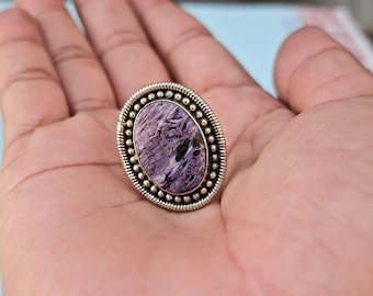 Op de afbeelding: Ovale zilveren ring met een grote, gepolijste charoietsteen met wervelende paarse en zwarte patronen. De steen is gezet in een kralenrand, wat een vintage-geïnspireerd ontwerp creëert. De ring is een opvallend sieraad.