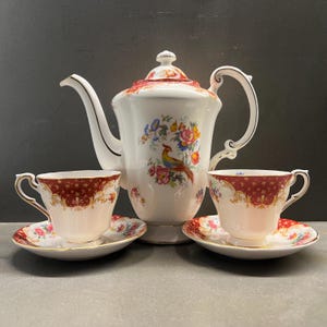 May include: A vintage tea set comprising a teapot and two teacups with saucers. The white porcelain teapot is adorned with a floral and bird motif, complemented by red accents. The teacups and saucers feature a red and gold pattern. Set on a grey surface.