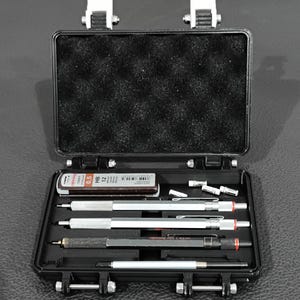 May include: An open black case containing various mechanical pencils, lead refills, and accessories. The case has a foam-lined top and secure latches. The pencils are silver and black with red accents. A small container of lead refills is also visible.